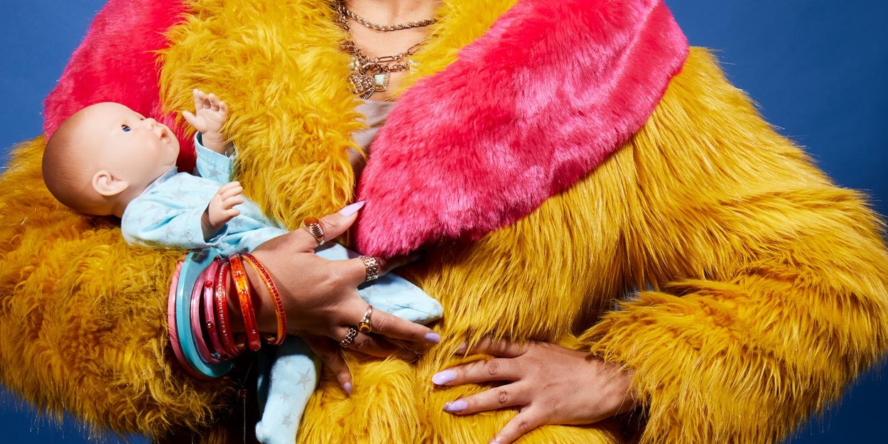 Baby Pearl Mackie Baby, starring Pearl Mackie, opens at the Brixton House in October 2026 (c) Clean House. Photo of a woman with the head cut off by the edge of the frame. She wears a yellow and pink fake fur coat and many cheap rings and necklaces. One hand is placed on her hip while the other hold a baby doll in a blue babygro.